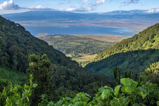 A scenic view of a lush valley surrounded by steep green hills, with a tranquil lake visible in the distance and mountains under a partly cloudy sky.